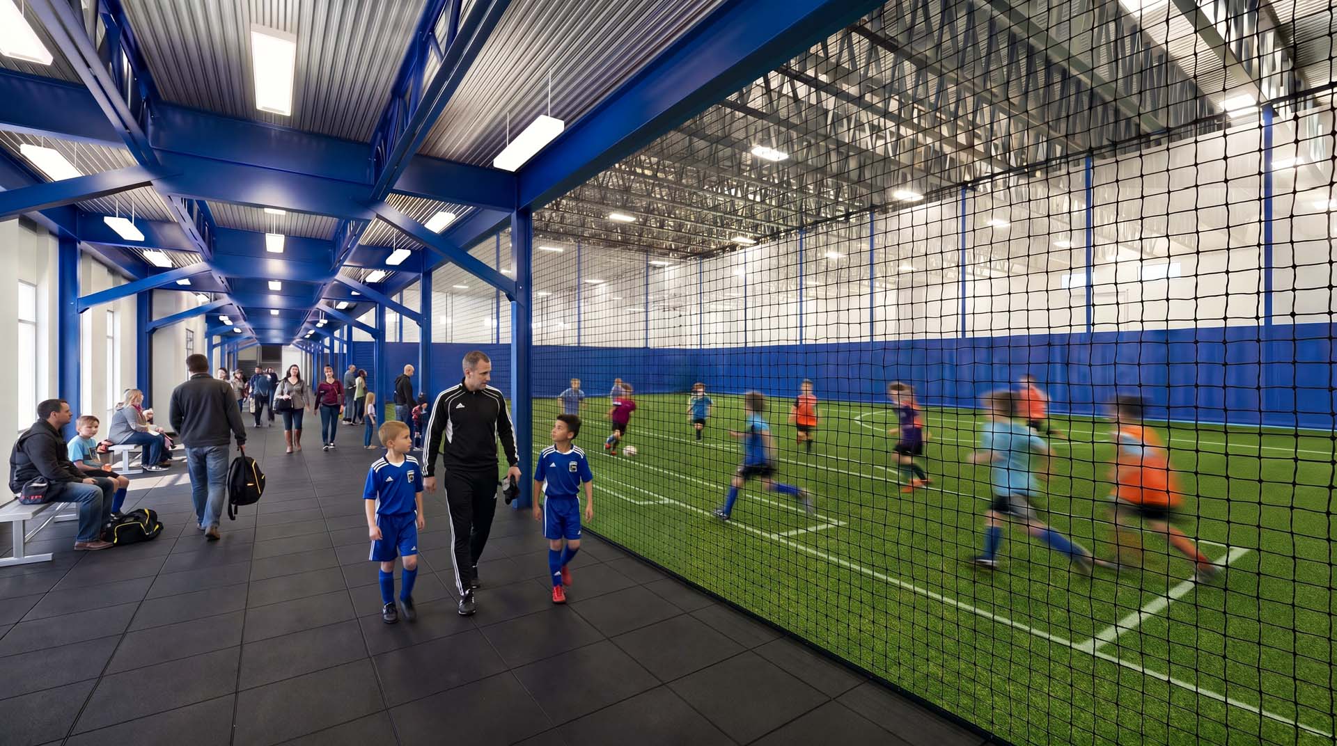 Indoor turf soccer field with youth players in team uniforms, viewed from a spectator walkway with families watching.