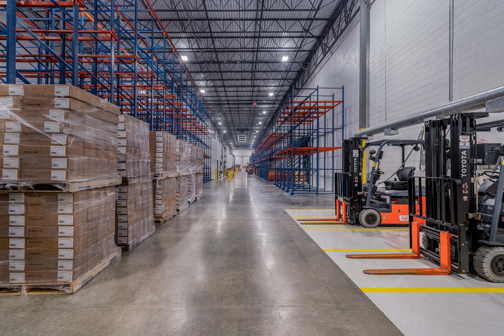 Warehouse aisle with palletized boxes, storage racks, and parked forklifts.