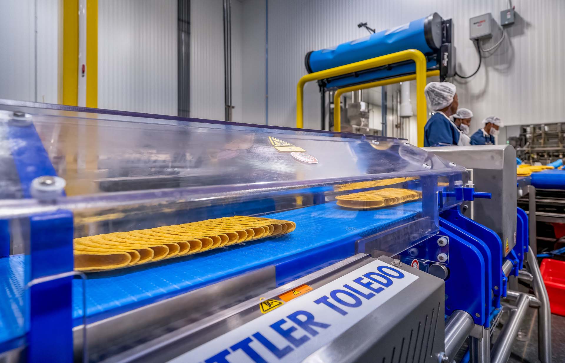 Tortillas on blue conveyor belt with workers in food production facility.