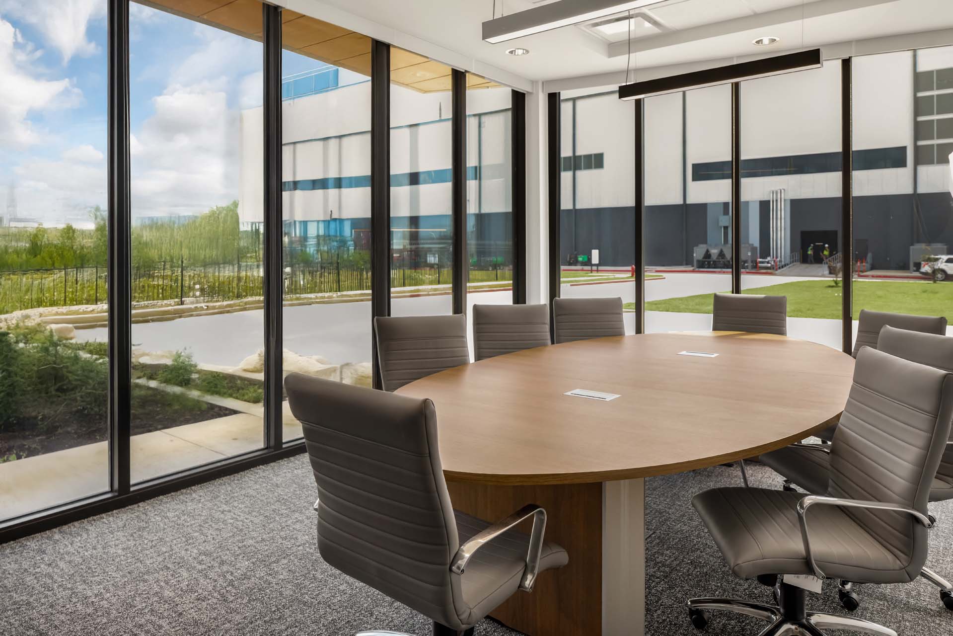Corner conference room with oval table, gray chairs, and floor-to-ceiling windows overlooking warehouse.