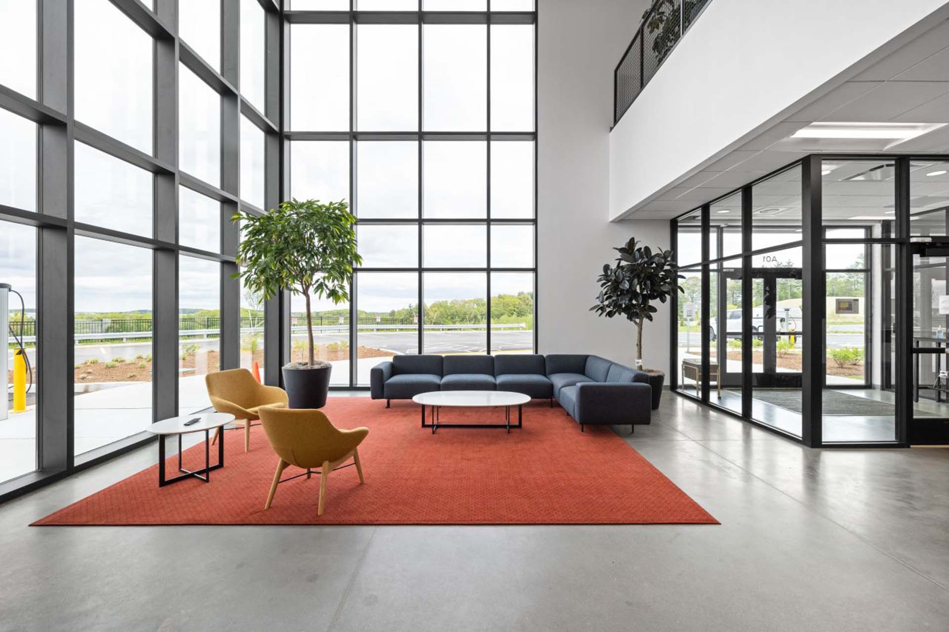 Two-story office lobby with floor-to-ceiling windows, navy sectional sofa, mustard chairs, and orange rug.