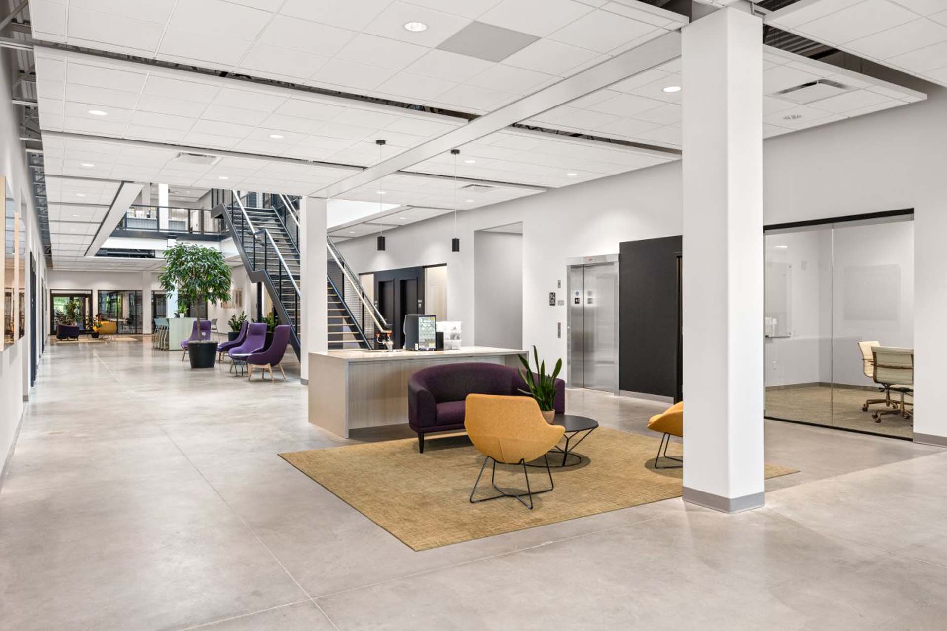 Modern office lobby with concrete floors, a metal staircase, purple and mustard lounge seating, a reception desk, and glass-walled conference room.