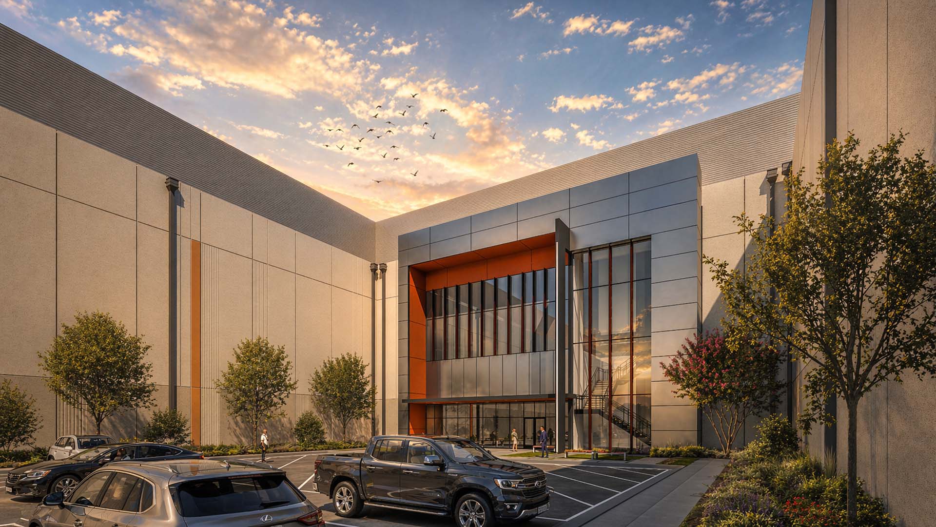 Industrial building entrance with glass curtain wall, orange accent panels, and parking lot at sunset.
